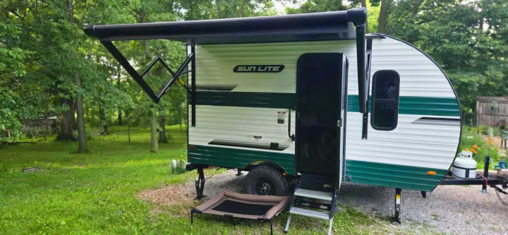 This Sun Lite RV rental features a cozy camper with an extended awning and pet bed, perfect for outdoor adventures in a wooded setting.. Sunset Park RV Sun Lite LTD 13MJ 2025
