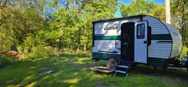 RV rental camping setup: Sun Lite travel trailer, steps & pet bed in a lush outdoor setting.. Sunset Park RV Sun Lite LTD 13MJ 2025