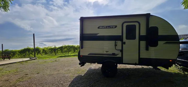 Cream & black Sun Lite RV rental parked at a sunny vineyard, featuring green vines, a wooden deck, and gravel ground.. Sunset Park RV Sun Lite LTD 13MJ 2025