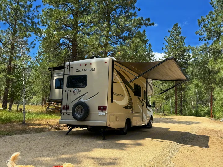 RV rental parked at a serene forest campsite with its awning extended under a bright blue sky, perfect for outdoor relaxation.. Thor Quantum 2018