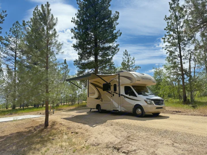 Scenic RV rental parked on a dirt path with its awning out, surrounded by tall pine trees under a bright blue sky.. Thor Quantum 2018