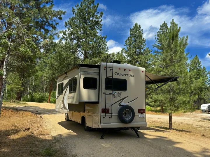 RV rental parked on a dirt road in a sunny forest campground, with its slide-out and awning extended.. Thor Quantum 2018