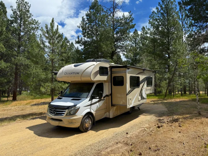 Scenic RV rental adventure! This motorhome is parked on a dirt road amidst tall pine trees under a cloudy blue sky, ready for a nature getaway.. Thor Quantum 2018
