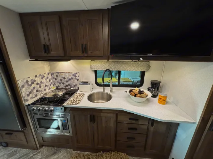Modern RV rental kitchen with dark wood cabinets, light counters, stove, microwave, sink, and mounted TV.. Forester LE 2022
