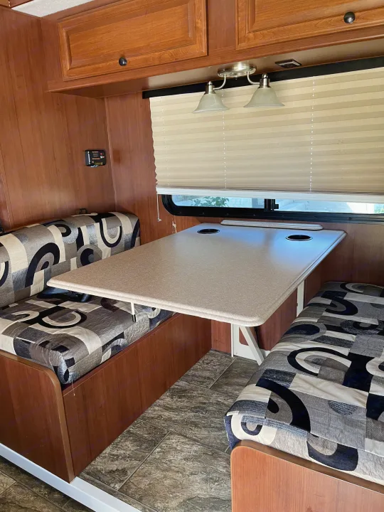 Inside an RV rental: a cozy dinette booth with patterned cushions, a table with cupholders, overhead storage, lights, and window blinds.. Tioga Montara 2012