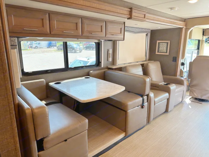Inside a modern RV rental: a light-toned dinette table, comfy recliners, and wood cabinets under large windows.. Thor Challenger 2022