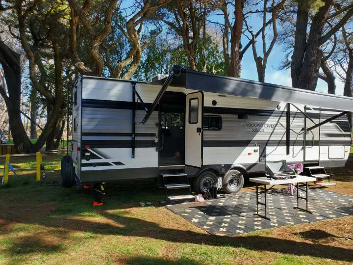 RV rental: a large travel trailer at a sunny, wooded campsite with awning, outdoor kitchen, and baby peeking out.. Grand Design Transcend Xplor 2021