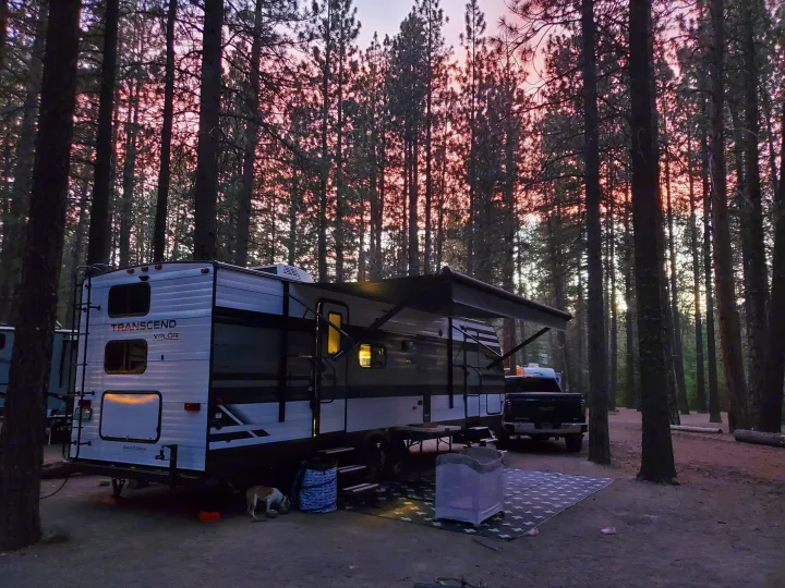 Sunset camping with an RV rental. A white Transcend Xplor travel trailer is parked in a pine forest, with a dog and mat outside.. Grand Design Transcend Xplor 2021