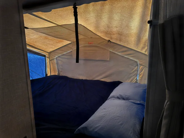 Inside a pop-up RV rental, a comfy bed with blue bedding and pillows awaits. Note the exit sign and ambient LED lighting.. Rockwood Roo 2022