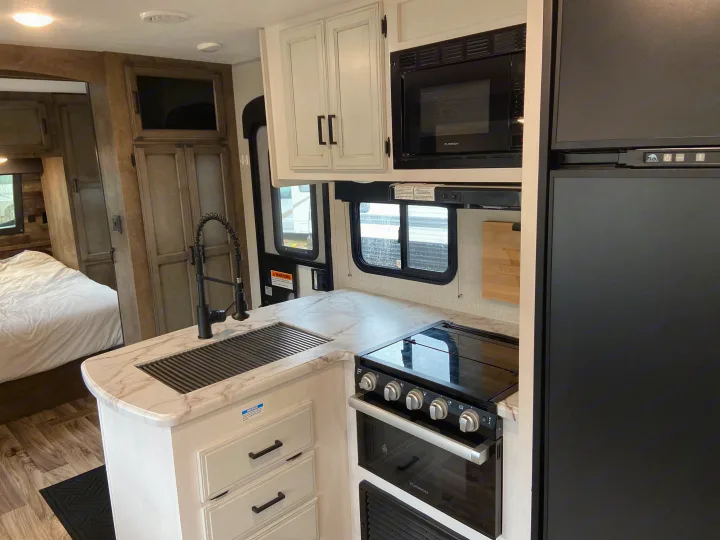Modern RV rental kitchen with white cabinets, marble counter, sink, stove, microwave, and fridge. Bedroom visible beyond.. Venture Sport Trek 2021