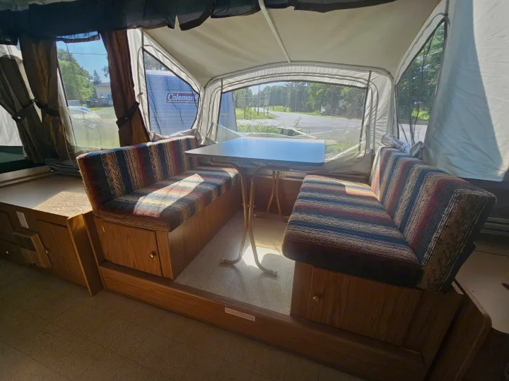 Step inside this cozy RV rental's pop-up camper dinette, featuring striped benches, a table, and bright window views.. Coleman Destiny 2000