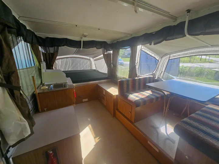 Inside a well-lit pop-up RV rental featuring a cozy bed, dinette with striped cushions, and compact kitchen counter.. Coleman Destiny 2000