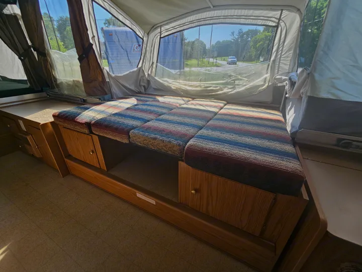 Inside a pop-up camper RV rental: striped bed, wood storage, and a bright view of trees & road.. Coleman Destiny 2000