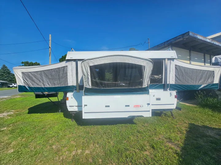 White pop-up RV rental, fully set up with mesh windows, parked on grass under clear sky. Ready for camping.. Coleman Destiny 2000