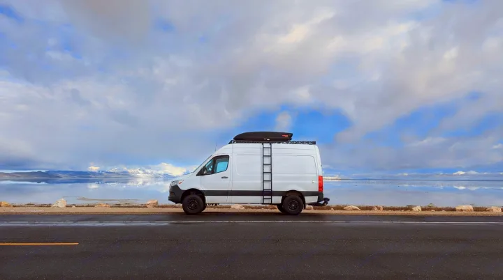 White Sprinter RV rental parked on a road beside a vast, reflective lake. Snow-capped mountains and cloudy skies in background.. Mercedes-Benz Sprinter 2019