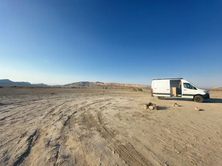 A white RV rental camper van parked in a wide-open, dry desert area with distant mesas. A rock fire pit is nearby.. Mercedes-Benz Sprinter 2019