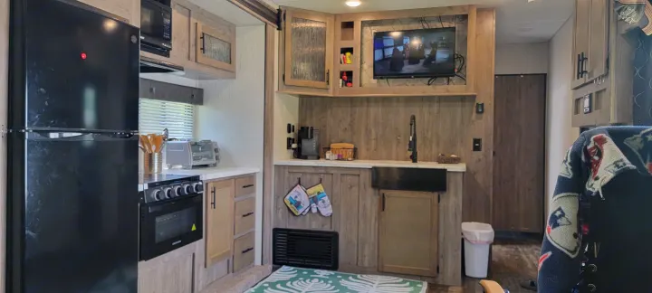 This RV rental features a modern kitchen area with a black fridge, stove, sink, and mounted TV showing a movie.. Forest River Cherokee Wolf Pack 2021