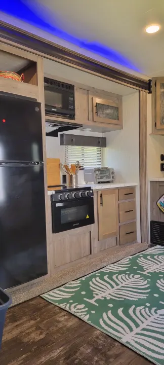 RV rental kitchen: black fridge, microwave, oven, light wood cabinets, and blue accent lighting, with a green patterned rug.. Forest River Cherokee Wolf Pack 2021