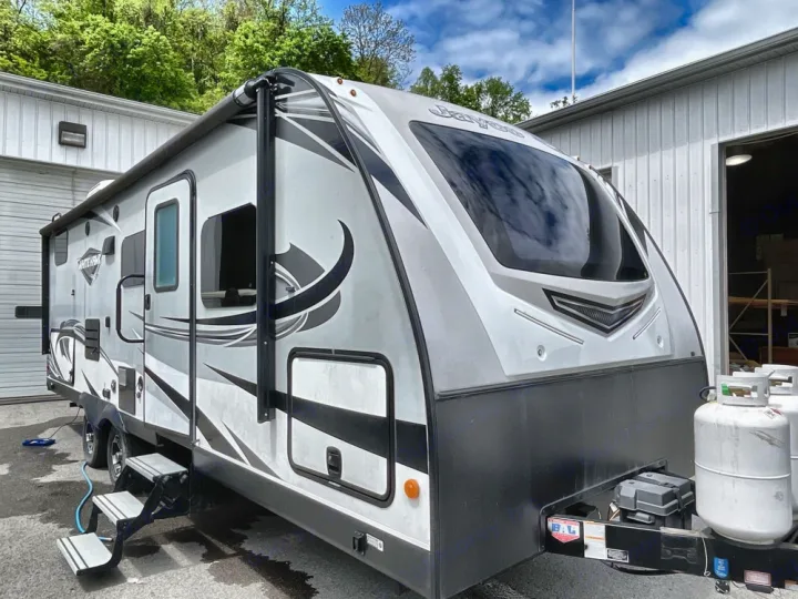Grey and black travel trailer parked outside, ready for your next RV rental. Features an awning, steps, and propane tanks for camping trips.. Jayco White Hawk 2019