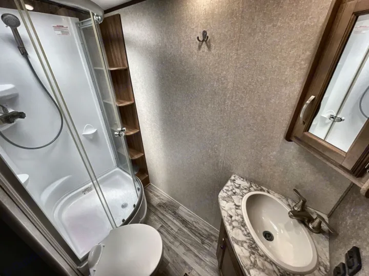 RV rental bathroom showcasing a compact corner shower, toilet, sink vanity, and built-in storage shelves.. Jayco White Hawk 2019