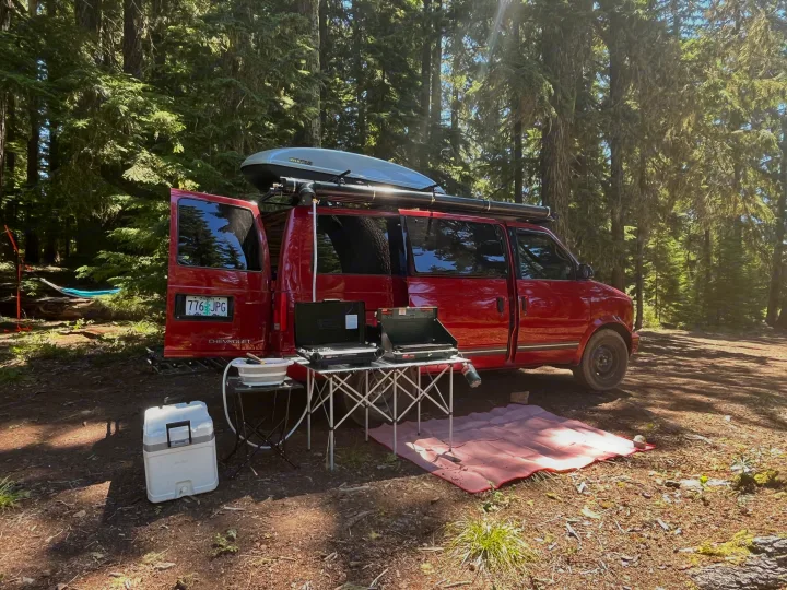 Red Chevy van, set up as an RV rental, camped in a sunny forest clearing with outdoor kitchen, hammock, and gear.. Chevrolet Astro Van 1997