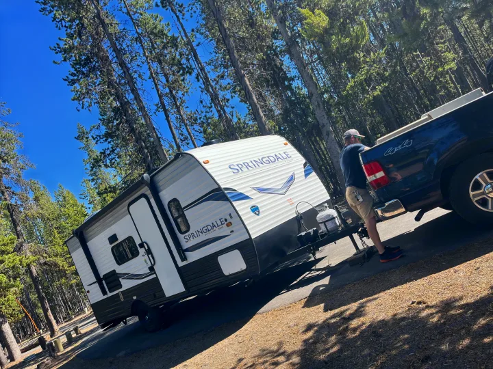 Man connecting an rv rental travel trailer to a truck under a clear blue sky among tall pine trees.. Keystone RV Springdale 2022