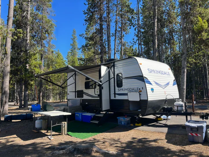 A white Springdale RV rental is set up in a sunny forest campsite. The travel trailer has its awning extended, showing a typical camping setup.. Keystone RV Springdale 2022