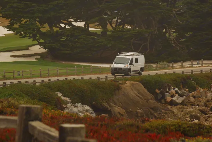 White RV rental van on a coastal road beside a green golf course, rugged shoreline, and gnarled trees.. Custom Ram Promaster 1500 2017