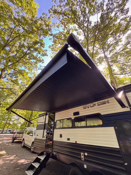 RV rental: A 'Cherokee Wolf Den' travel trailer with its awning extended, parked at a sunny, tree-lined campsite.. Forest River Cherokee Grey Wolf 2024