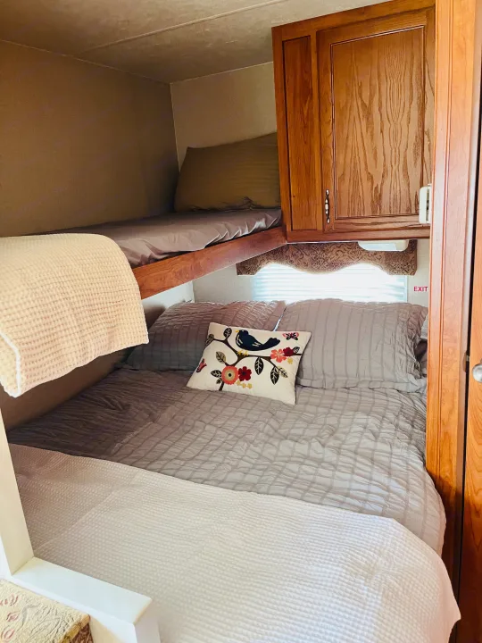 Cozy RV rental interior featuring a lower bed with gray bedding, an upper bunk bed, and wooden cabinets. An "EXIT" sign is visible.. Dutchmen Rainier 2006