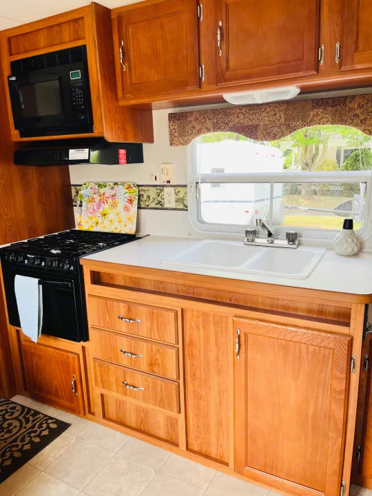 Cozy RV rental kitchen: black microwave, stove, double sink, light wood cabinets, and a window view.. Dutchmen Rainier 2006