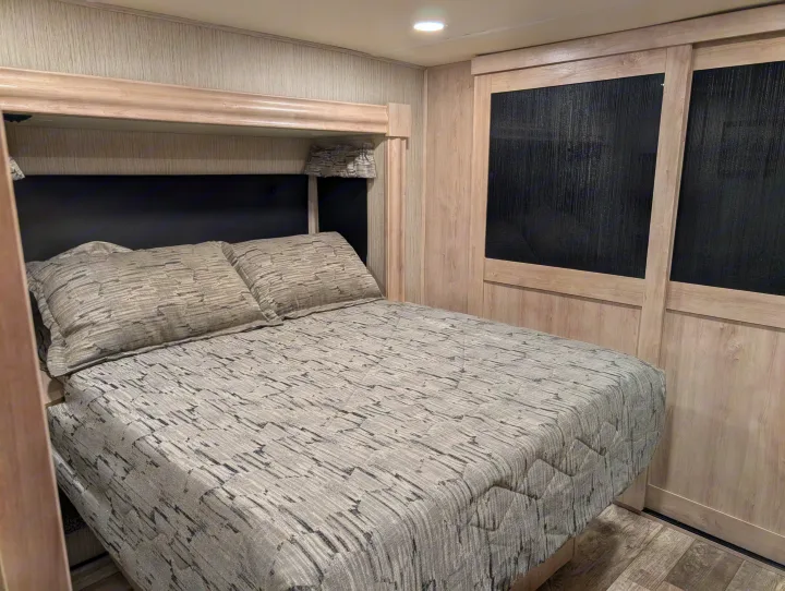 RV rental bedroom: a neat space with grey patterned bedding, black headboard, and light wood walls/cabinets.. Forest River Sandpiper 2022