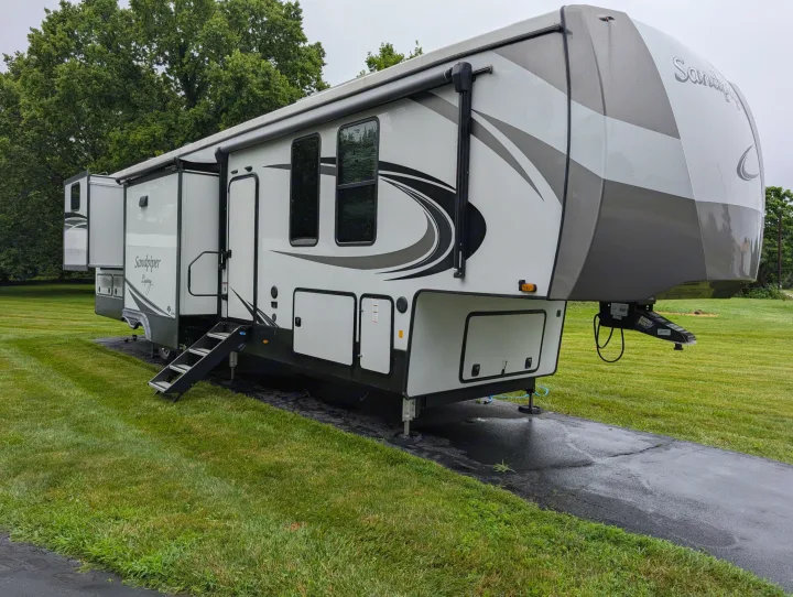 This luxury Sandpiper fifth-wheel RV rental is parked on a wet driveway surrounded by green grass.. Forest River Sandpiper 2022
