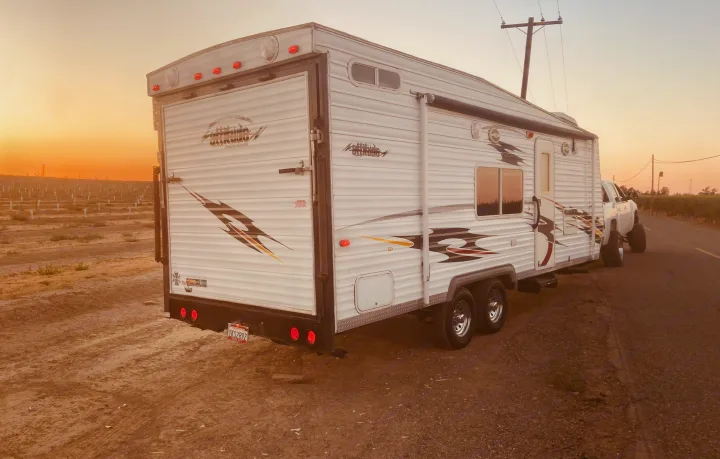 A white RV rental toy hauler is parked on a rural dirt road next to an orchard at sunset, hitched to a pickup truck.. Eclipse Recreational Vehicles Attitude 2006