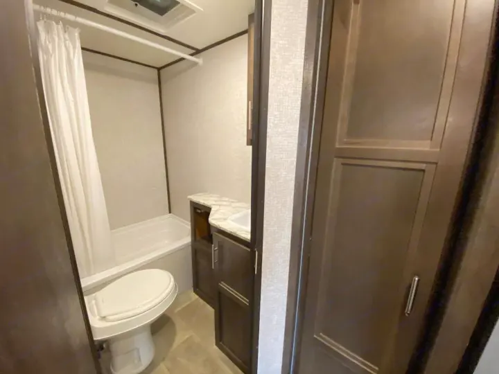 Inside an RV rental bathroom: a white toilet, compact tub/shower with curtain, and dark wood vanity with sink. Practical for travel.. Coleman 2125bh 2019