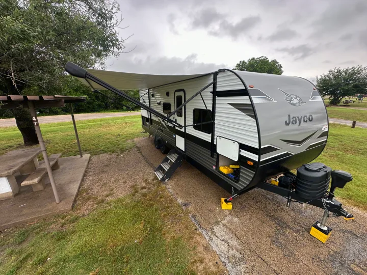 Grey Jayco travel trailer at a cloudy campsite, ready for an RV rental adventure. Awning extended, with a picnic table.. Jayco Jay Flight 2024