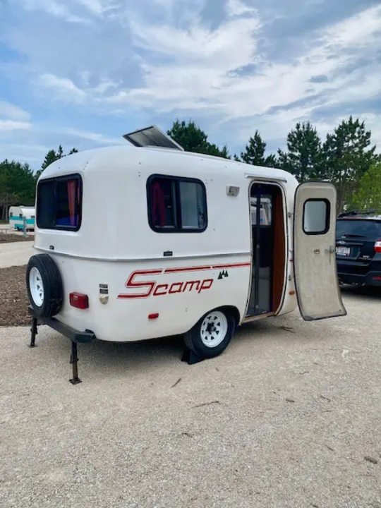 Explore the outdoors! This white Scamp travel trailer, an excellent RV rental, awaits at a gravel campsite.. Scamp Scamp Trailer 1999