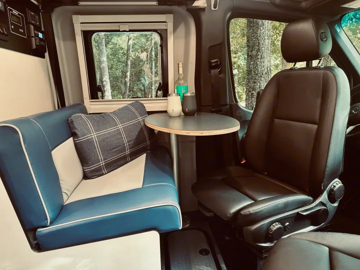 Step inside this comfortable RV rental. Features a blue bench seat, round table with drinks, and a serene forest view.. Winnebago Revel 2025
