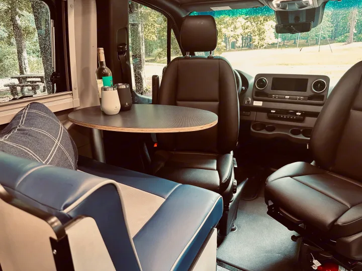 Cozy RV rental interior with blue seating, a table with drinks, and comfortable front seats, viewing a rainy park.. Winnebago Revel 2025