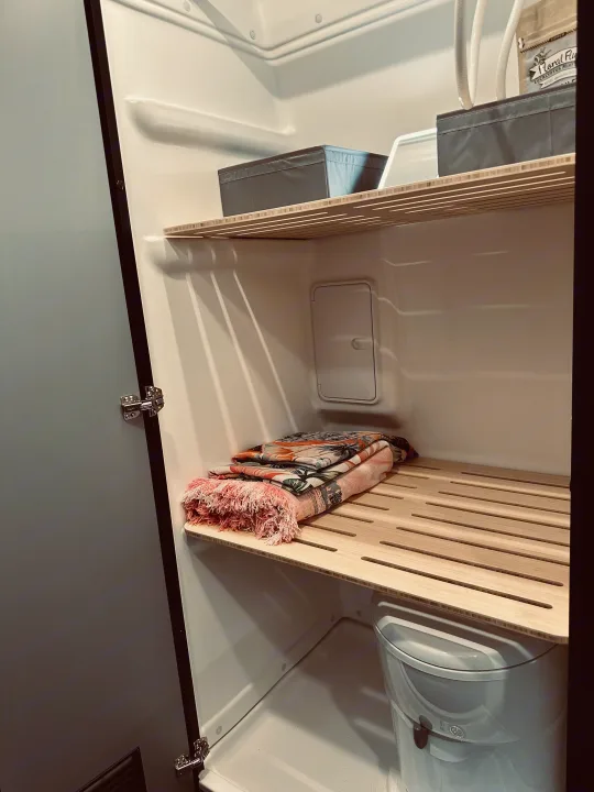 Inside an RV rental's compact bathroom, showing slatted wooden shelves with storage bins, folded blankets, and a toilet.. Winnebago Revel 2025