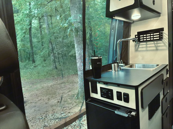 Inside an RV rental: a kitchenette with sink, controls, and fridge, looking out to a serene forest scene through a screen. Perfect for camping!. Winnebago Revel 2025