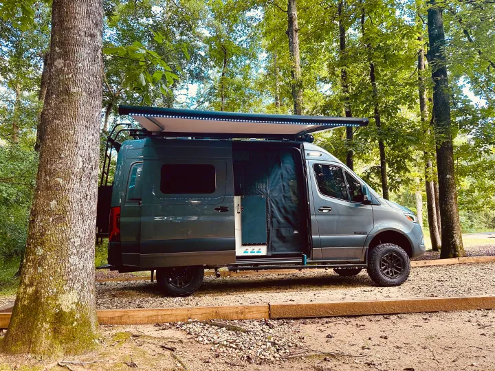 Your next outdoor adventure starts with this Winnebago RV rental. A grey camper van with an illuminated awning, parked in a wooded campsite.. Winnebago Revel 2025