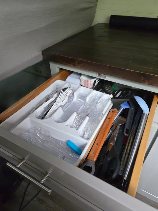 Inside an RV rental drawer: a white tray holds clear plastic cutlery, alongside spatulas, tongs, and other cooking tools. Essential camping gear.. Freightliner Sprinter 2011