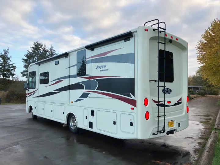 White Jayco Precept RV rental, a large motorhome with a ladder, parked on wet pavement under a cloudy sky.. Jayco Precept 2018