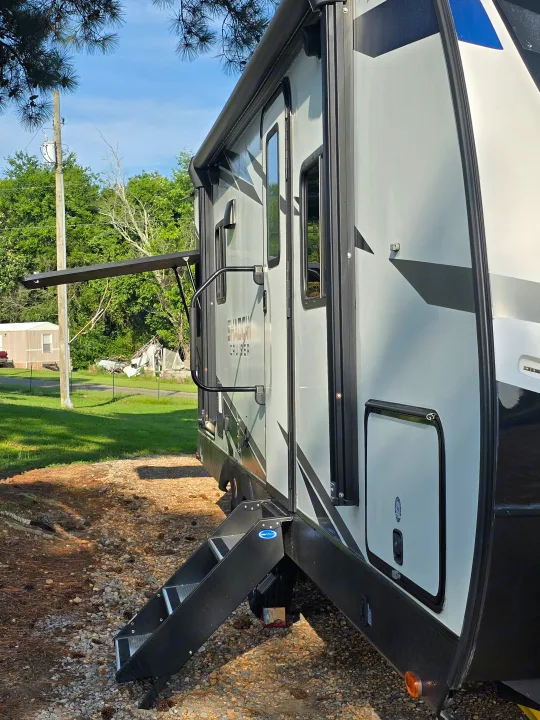 Side of a light-colored RV rental with a black awning extended and steps down, parked on a gravel lot under blue sky.. Cruiser RV Shadow Cruiser 2021