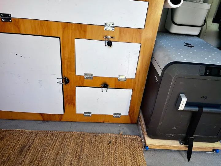 An rv rental's interior: custom wood cabinetry with white doors, a large Dometic CFX cooler, and a woven mat.. Dodge Ram Promaster 2500 2021