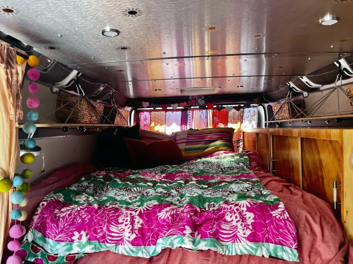 Inside a vibrant RV rental: cozy bed with pink/green quilt, wooden cabinets, and string lights create a warm, inviting space.. Dodge Ram Promaster 2500 2021
