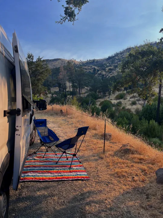 An rv rental set up for camping: white camper van, blue chairs, and a colorful mat overlook a dry grassy hillside.. Dodge Ram Promaster 2500 2021