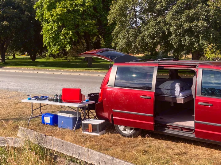 This red minivan camper conversion, like an RV rental, is set up in a park with a cooking table and a bed.. Dodge Grand Caravan 2010