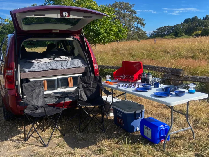 An RV rental-style setup: Minivan with bed, outdoor kitchen & cooking supplies, and chairs in a sunny field.. Dodge Grand Caravan 2010
